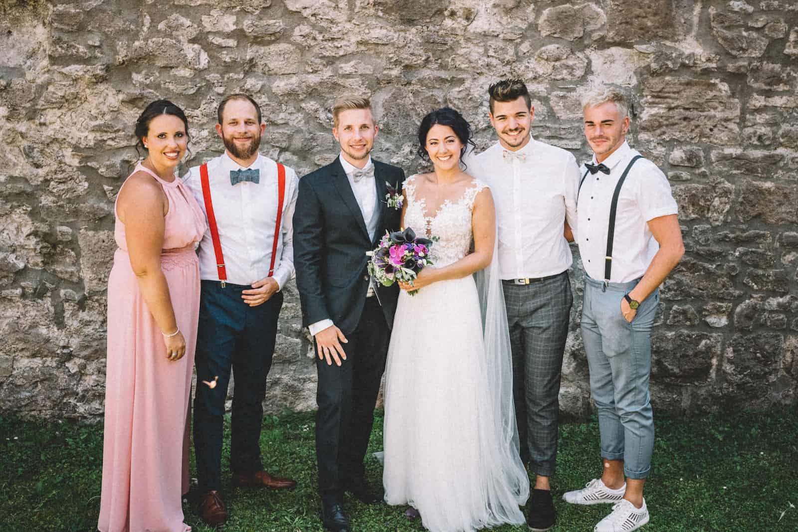 Hochzeitsfotografie in Wüstenrot mit Brautpaar und Gästen vor einer Steinmauer, aufgenommen vom Hochzeitsfotografen Niklas Hesser.