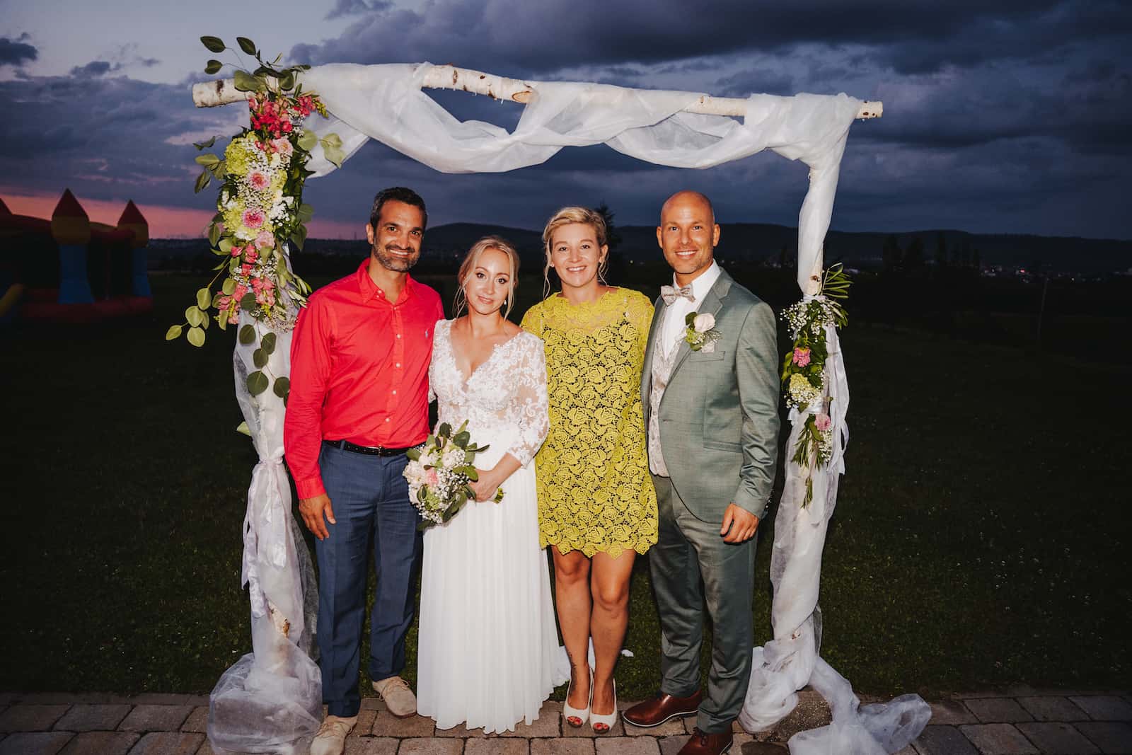 Hochzeitsfotografie in Kernen mit Brautpaar und Gästen unter einem dekorierten Bogen bei Abenddämmerung.