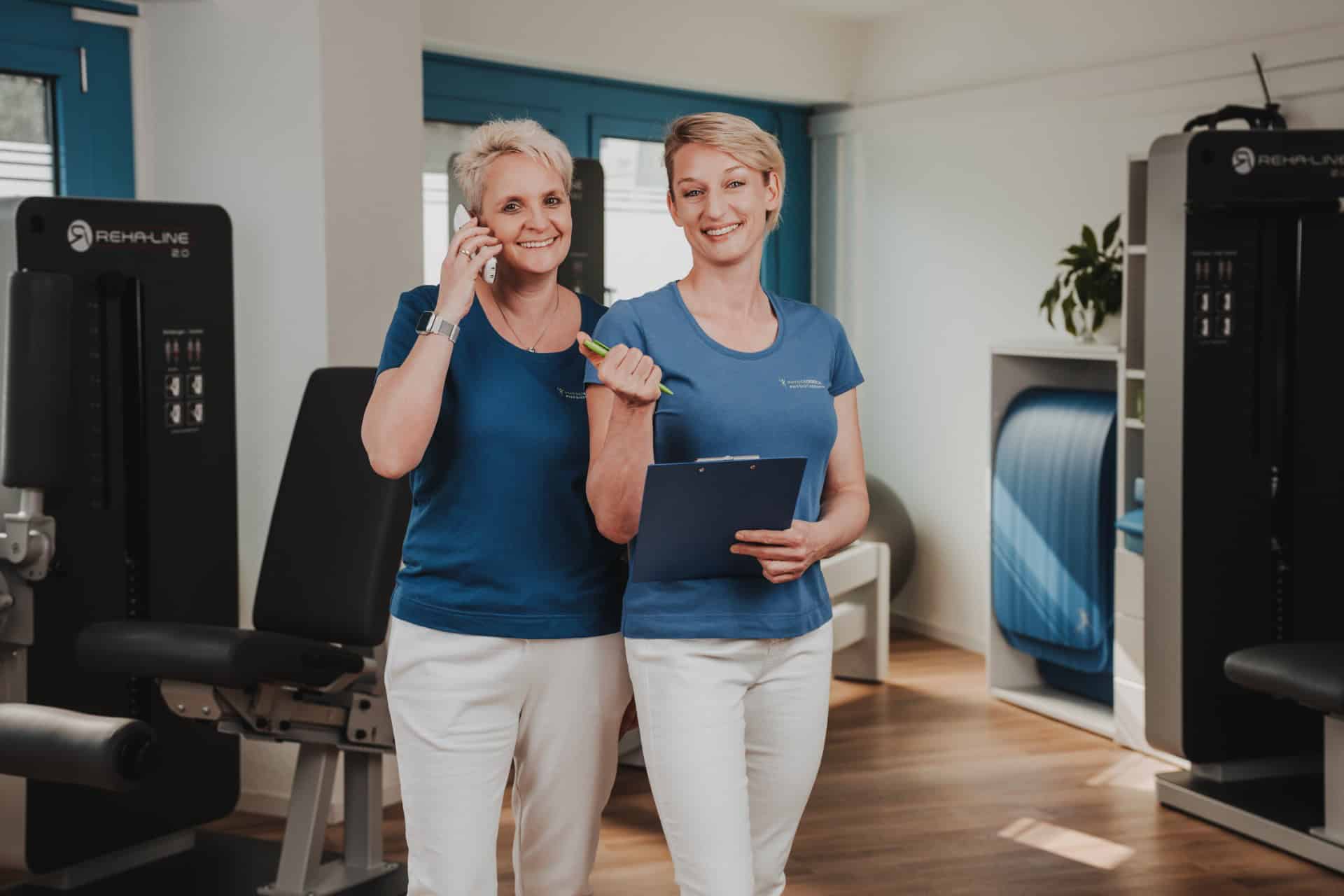 Die Inhaberinnen von PhysioLogisch Erlenbach im Praxisalltag – eine telefoniert, die andere hält Klemmbrett, umgeben von REHA-LINE Geräten