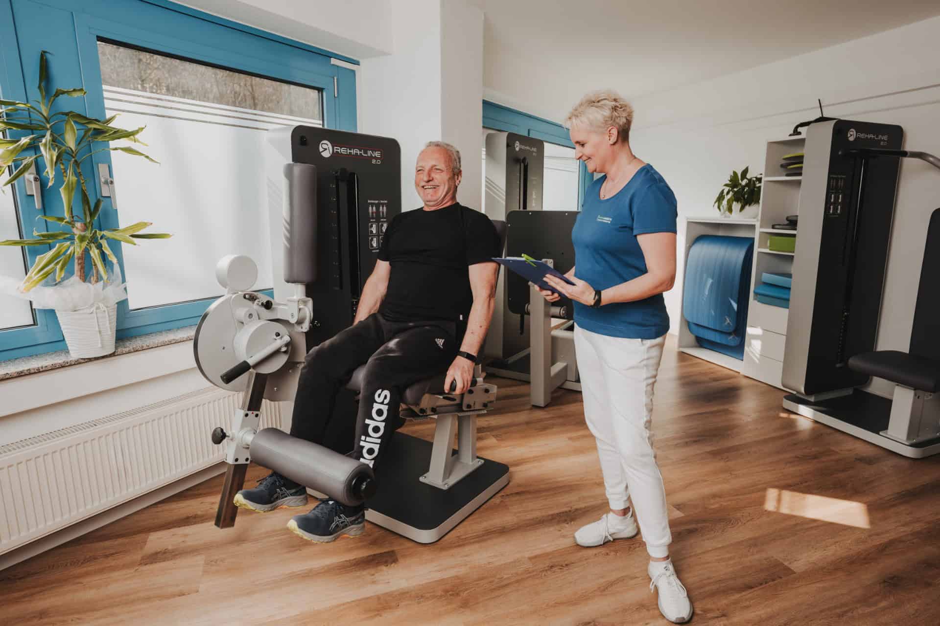 Physiotherapeutin von PhysioLogisch Erlenbach im freundlichen Gespräch mit älterem Patienten beim Reha-Training an der REHA-LINE 2.0