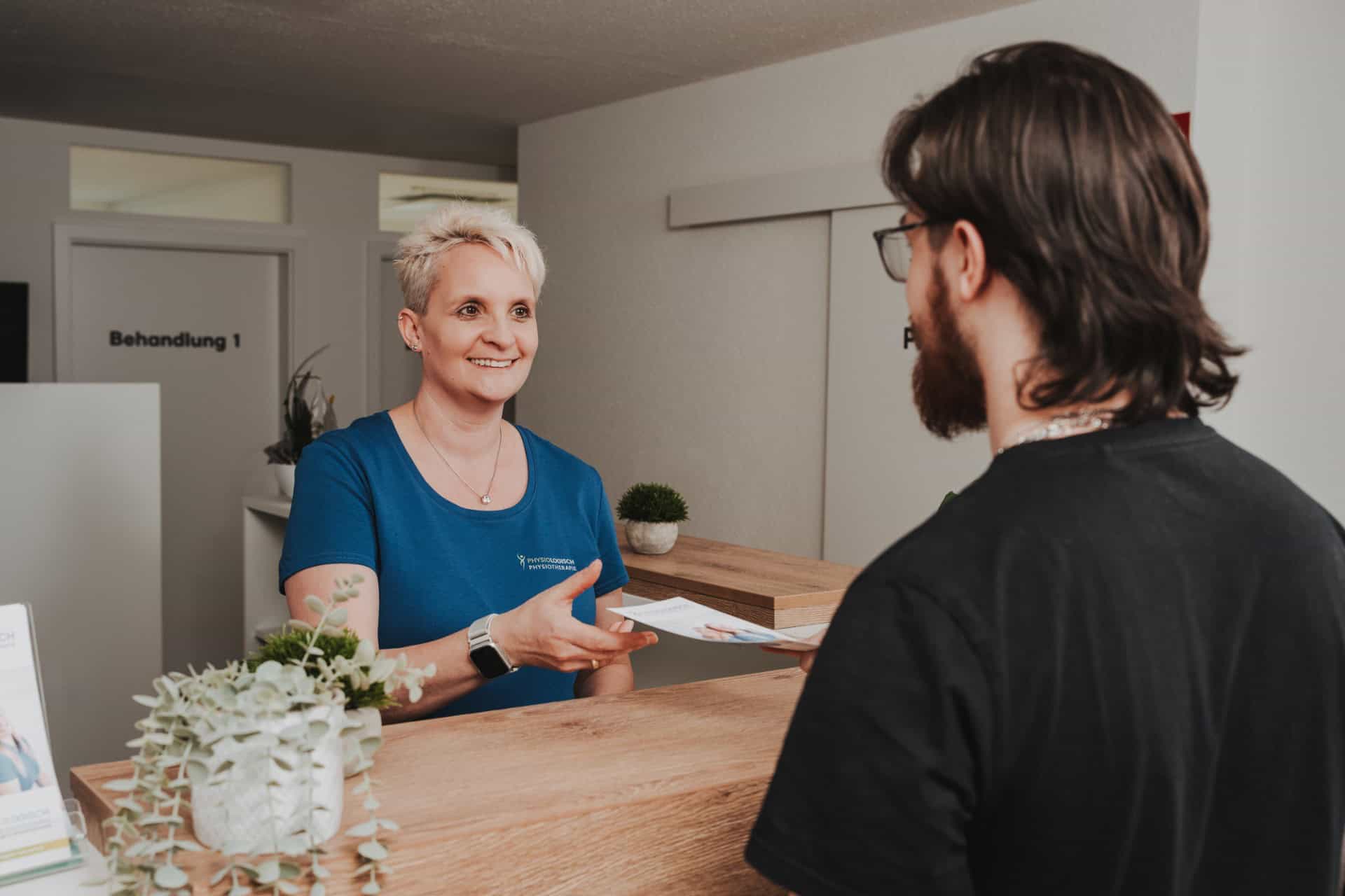 Mitarbeiterin von PhysioLogisch Erlenbach überreicht einem Patienten an der Rezeption Unterlagen im Empfangsbereich der Praxis