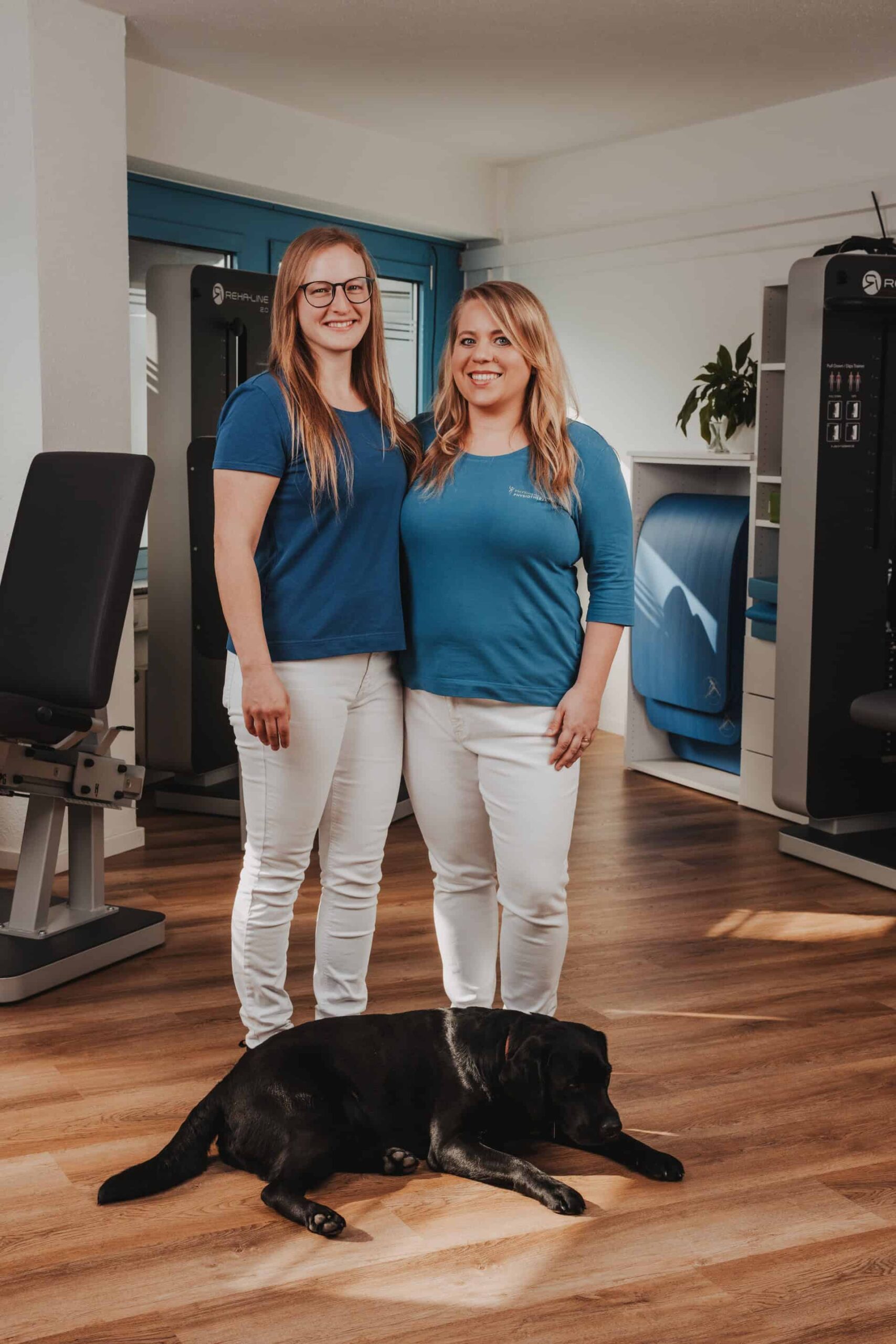 Zwei Physiotherapeutinnen von PhysioLogisch Erlenbach lächeln mit ihrem schwarzen Labrador im modern ausgestatteten Praxisraum