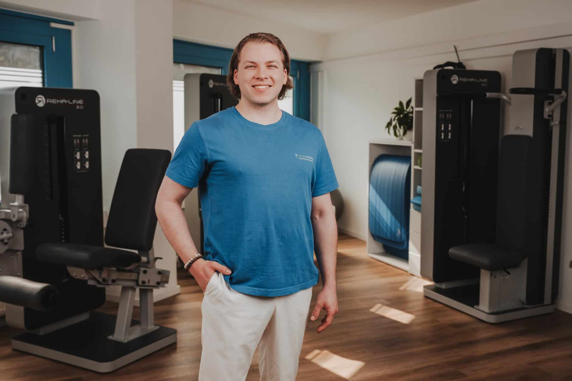Freundliches Portrait eines Therapeuten von PhysioLogisch Erlenbach im blauen Praxis-Shirt vor modernen REHA-LINE Trainingsgeräten