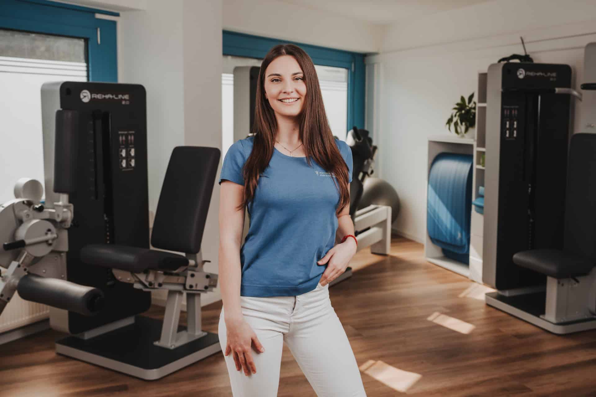 Portrait einer Physiotherapeutin von PhysioLogisch Erlenbach mit langen dunklen Haaren im blauen Praxis-Shirt vor modernen Trainingsgeräten