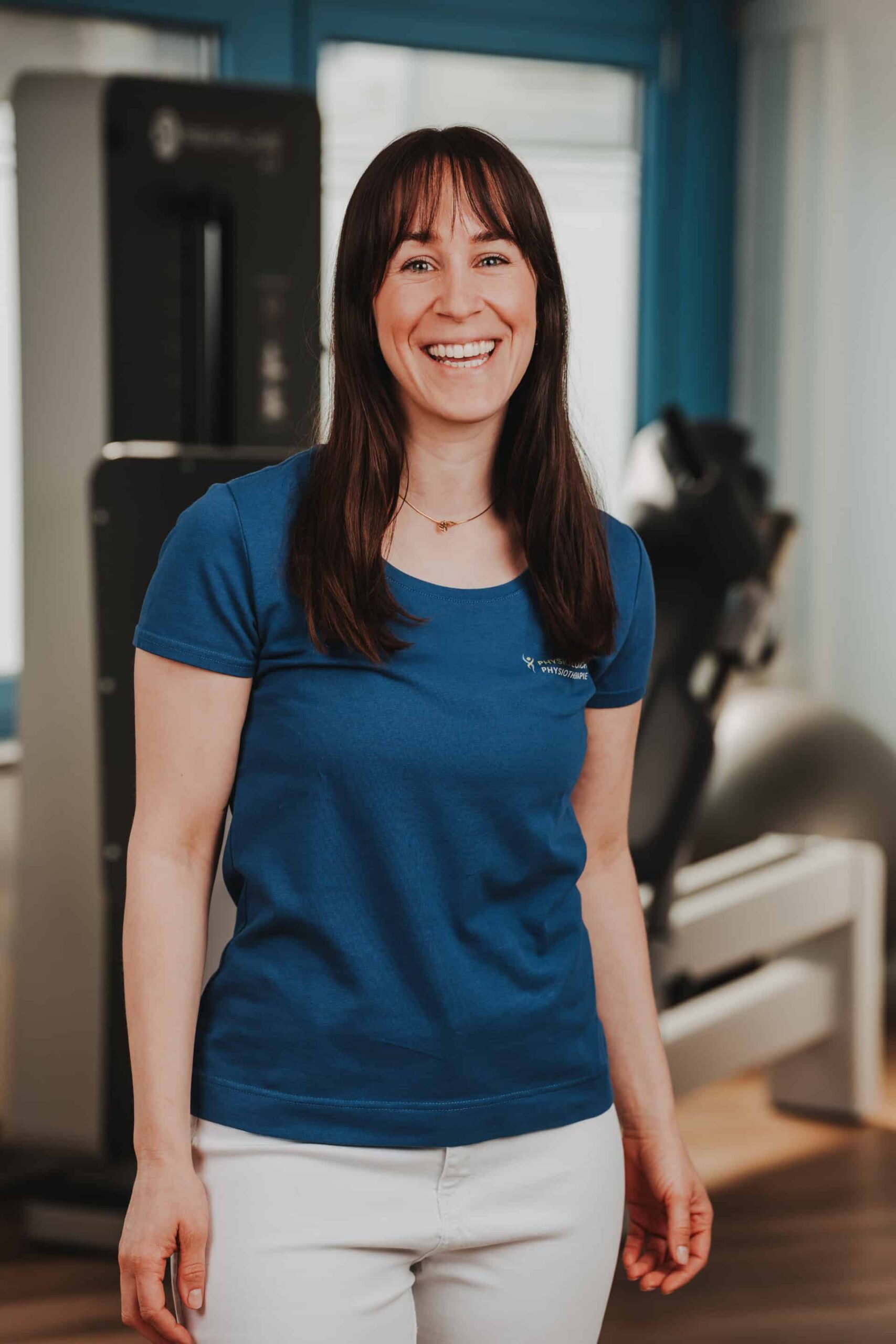 Freundliches Portrait einer Physiotherapeutin von PhysioLogisch Erlenbach im blauen Praxis-Shirt vor dem Geräteraum der Praxis