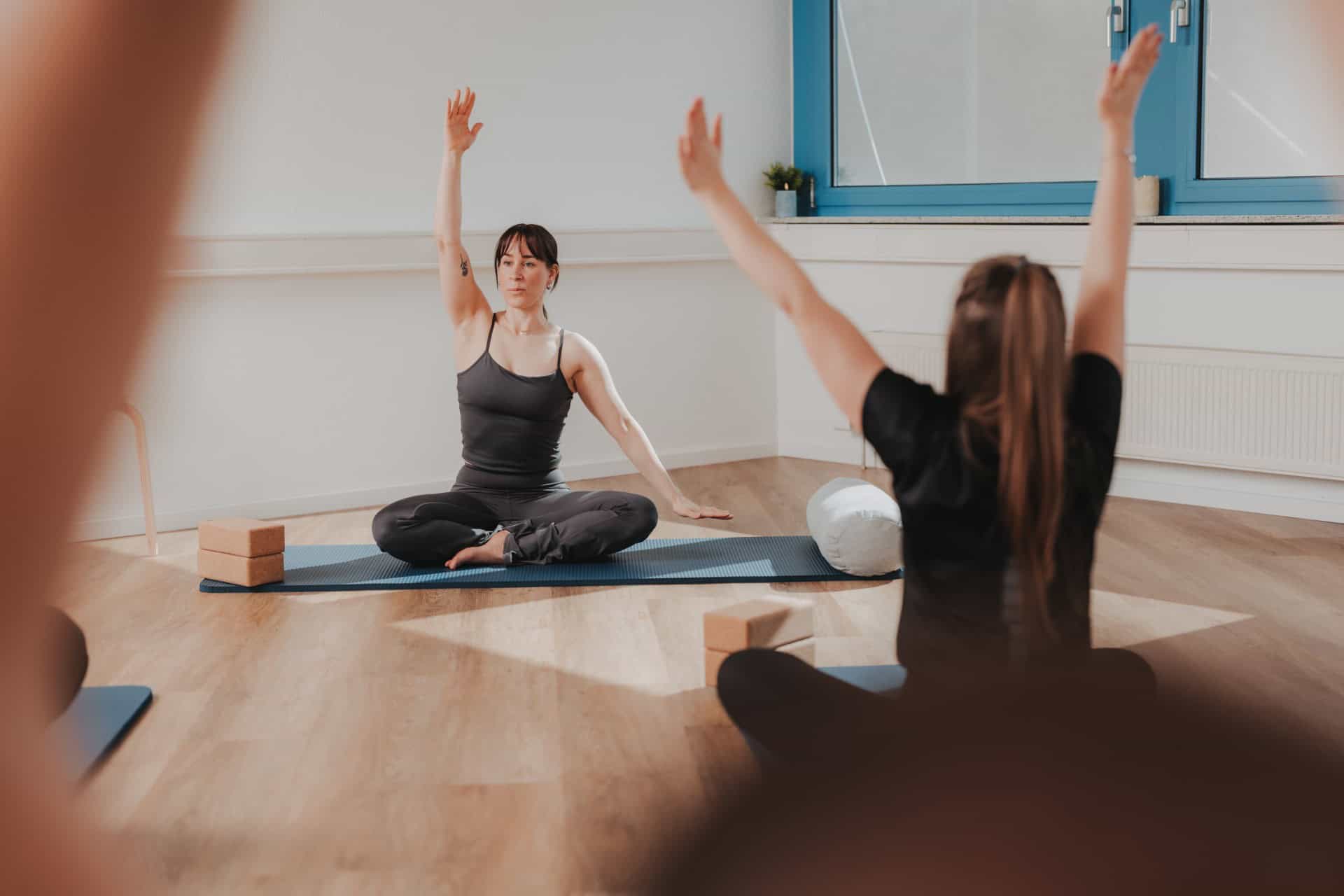 Yoga-Kurs bei PhysioLogisch Erlenbach – Kursleiterin zeigt Dehnübung im Schneidersitz mit erhobenem Arm, Teilnehmerin folgt der Bewegung